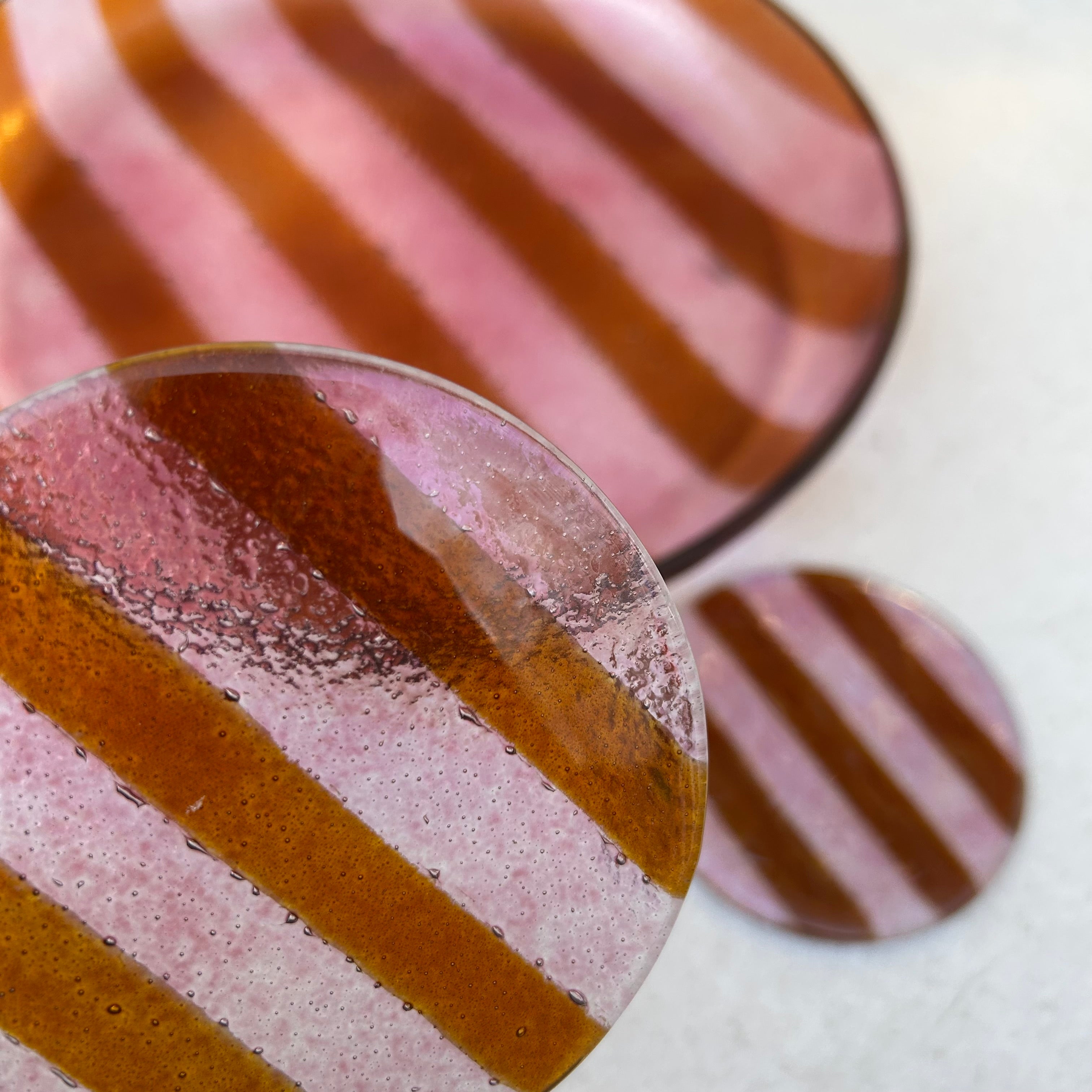 Sfeerbeeld verschillende glazen borrelitems. Close up van glazen ronde onderzetter met roze strepen met op de achtergrond nog een onderzetter en een glazen ronde serveerschaal met roze strepen.