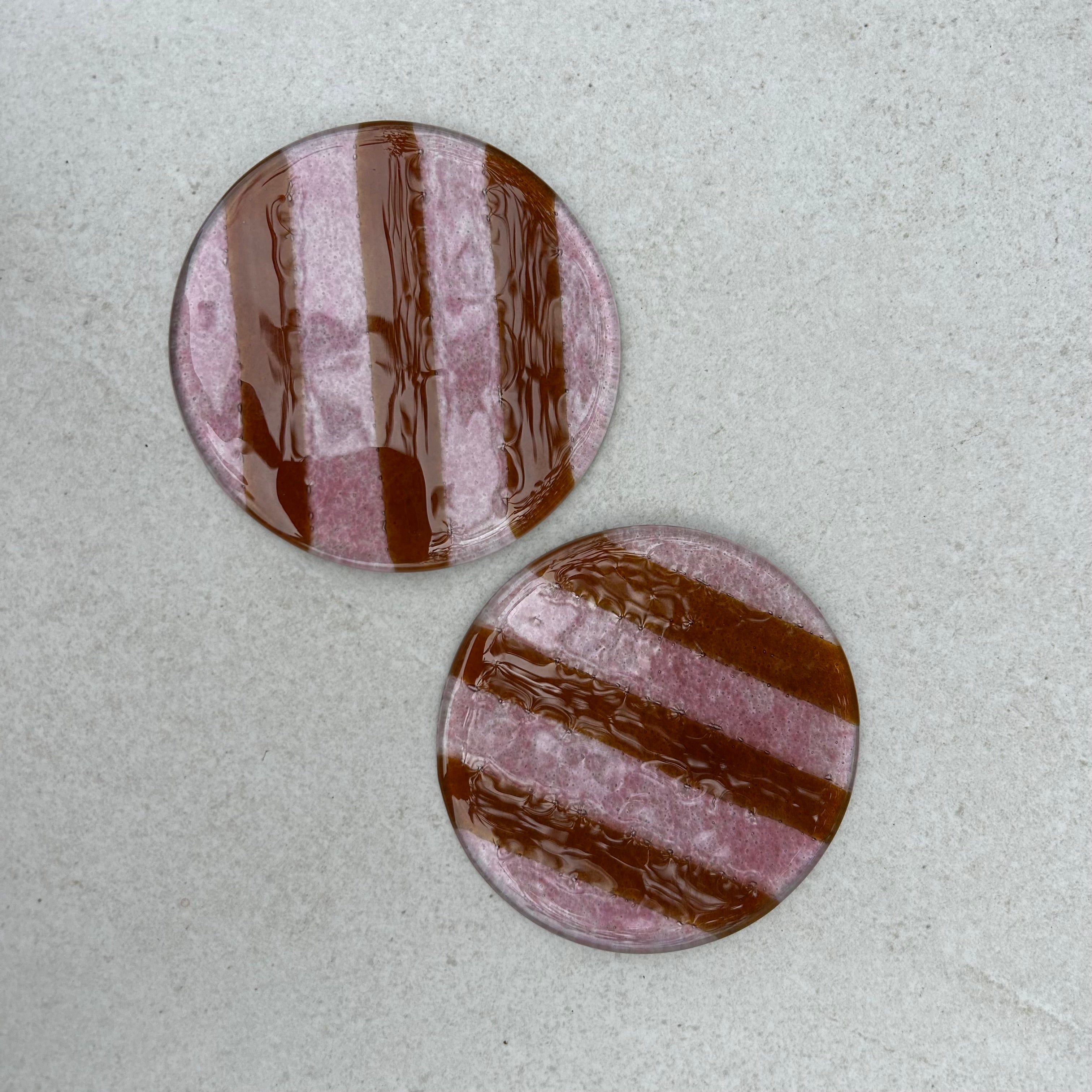 Bovenaanzicht twee glazen ronde onderzetters met roze strepen op een beige ondergrond.