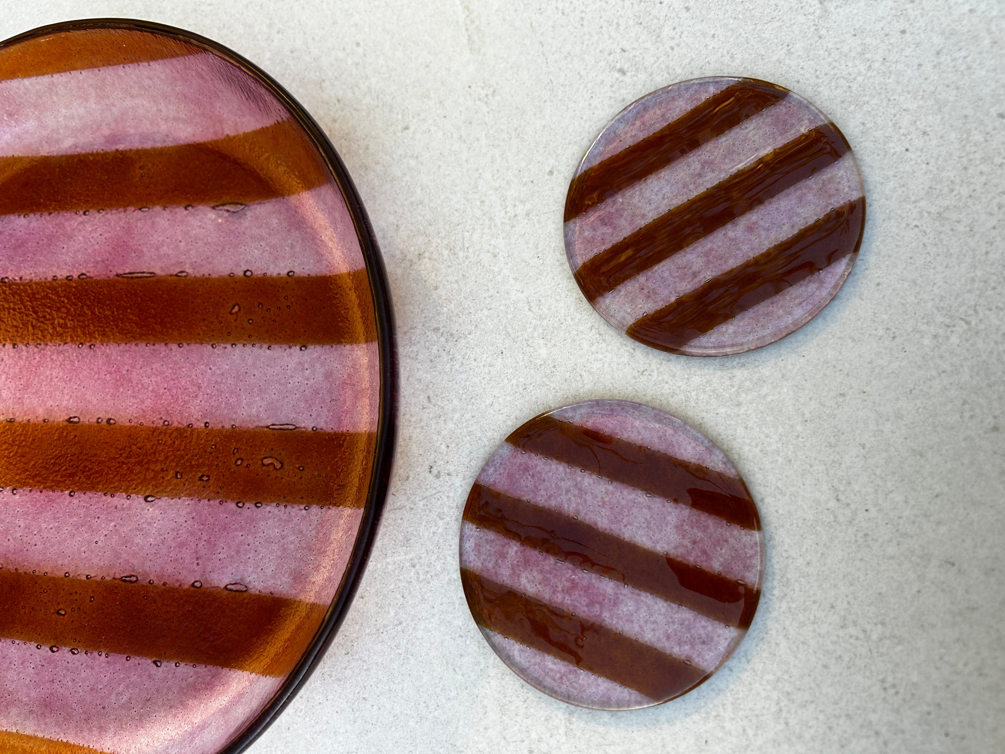 Sfeerbeeld van twee glazen ronde onderzetters met roze strepen en een grote glazen ronde serveerschaal met roze strepen op een beige achtergrond.