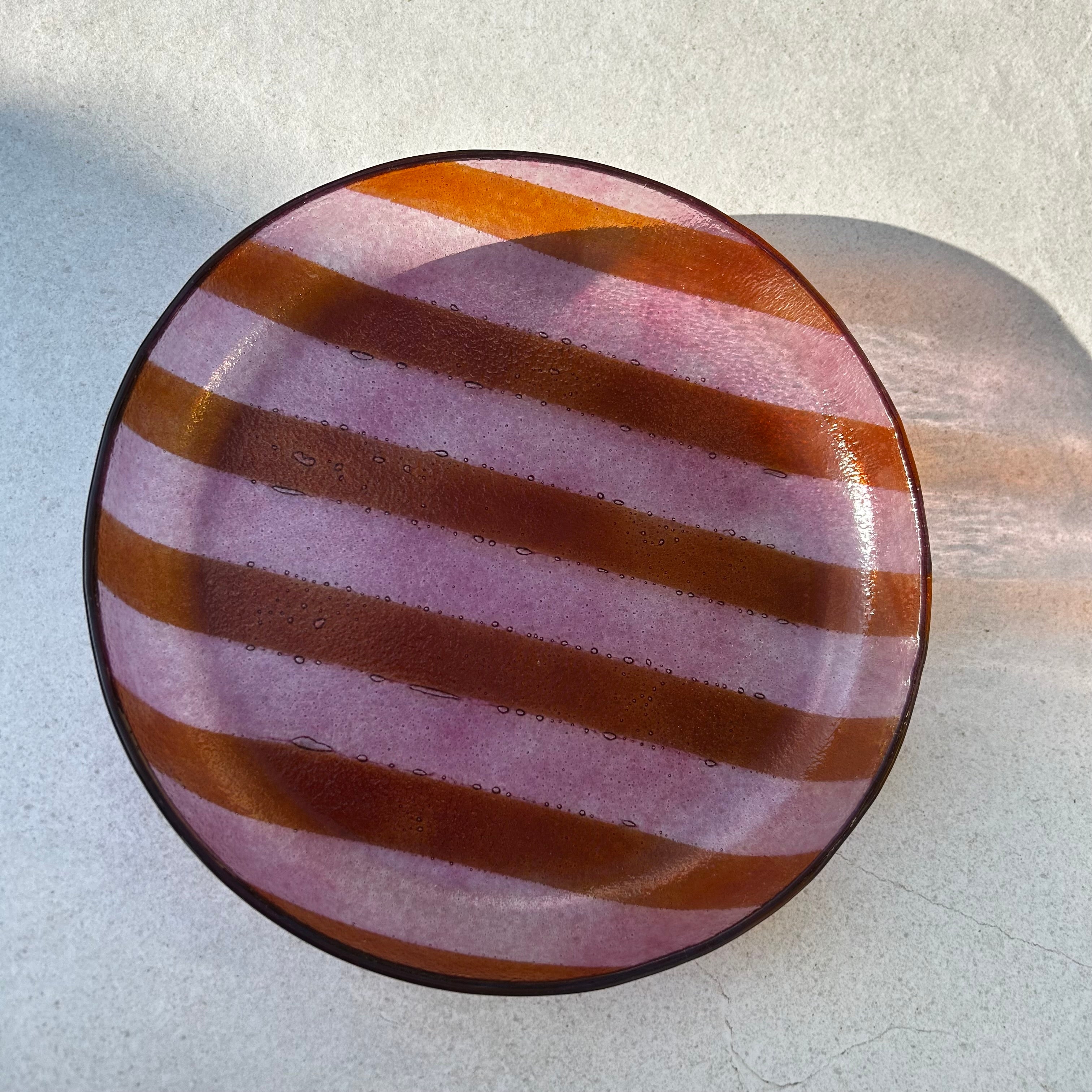Bovenaanzicht van een ronde glazen serveerschaal met oranje en roze strepen op een beige achtergrond.