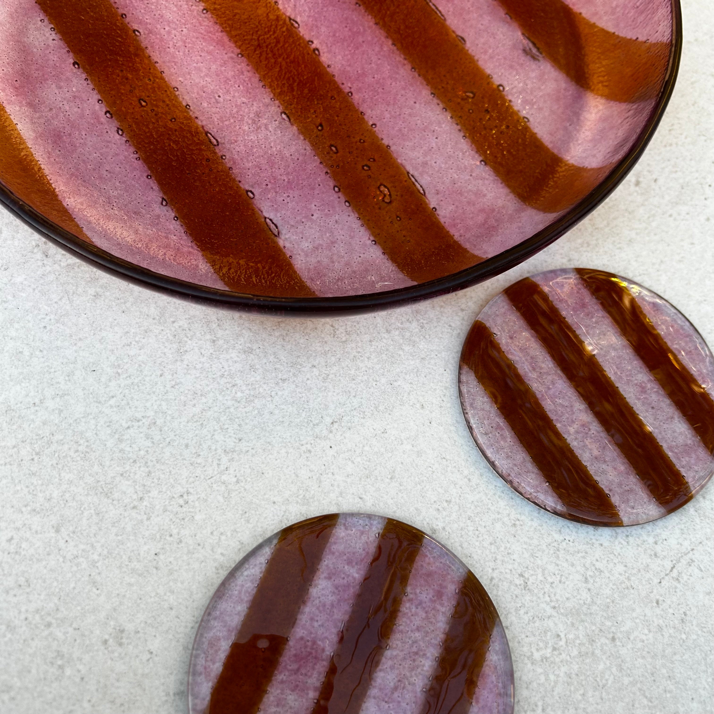 Bovenaanzicht van een ronde roze glazen serveerschaal met oranje strepen en 2 ronde glazen onderzetters, roze met oranje strepen op een witte achtergrond.